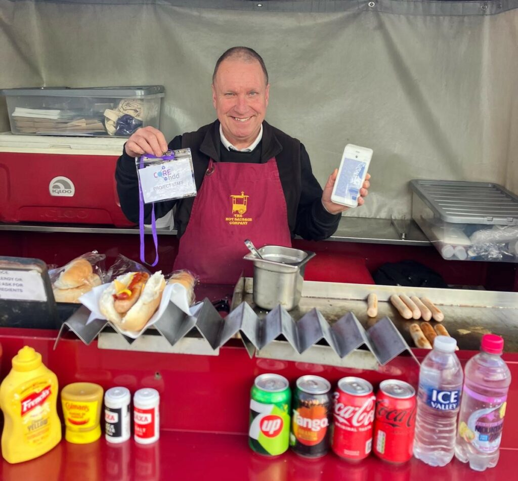 Andy at the Hot Sausage Co kiosk holding up a connected device. 